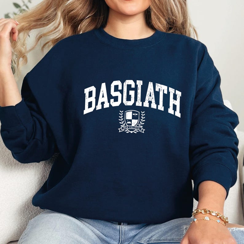 以下が含まれることがあります： 紺色のスウェットシャツ。白いブロック体で「BASGIATH」の文字がプリントされています。テキストの下には紋章のデザインがあります。クルーネックのスタイルです。