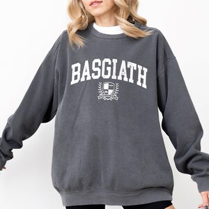以下が含まれることがあります： 胸に白い文字で「BASGIATH」と書かれたチャコールグレーのスウェットシャツ。テキストの下には紋章のデザインがあります。スウェットシャツはクルーネックと長袖です。