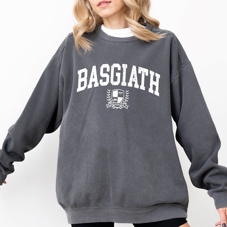 以下が含まれることがあります： 胸に白い文字で「BASGIATH」と書かれたチャコールグレーのスウェットシャツ。テキストの下には紋章のデザインがあります。スウェットシャツはクルーネックと長袖です。