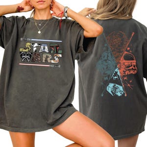 K&ouml;nnte beinhalten: Dunkelgraues T-Shirt mit dem Text "STAR WARS" in einem farbenfrohen Blockbuchstaben-Design. Die Vorderseite des Shirts zeigt Lichtschwerter und Charakterbilder. Die R&uuml;ckseite des Shirts zeigt einen Todesstern, Darth Vader, einen Sturmtruppler und TIE-J&auml;ger.