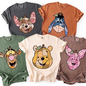 May include: Five short-sleeved t-shirts in various colors, each featuring a cartoon character's face. The characters include Eeyore, Piglet, Tigger, and Winnie the Pooh, with a pumpkin-like design. The shirts are in shades of gray, orange, green, cream, and brown.