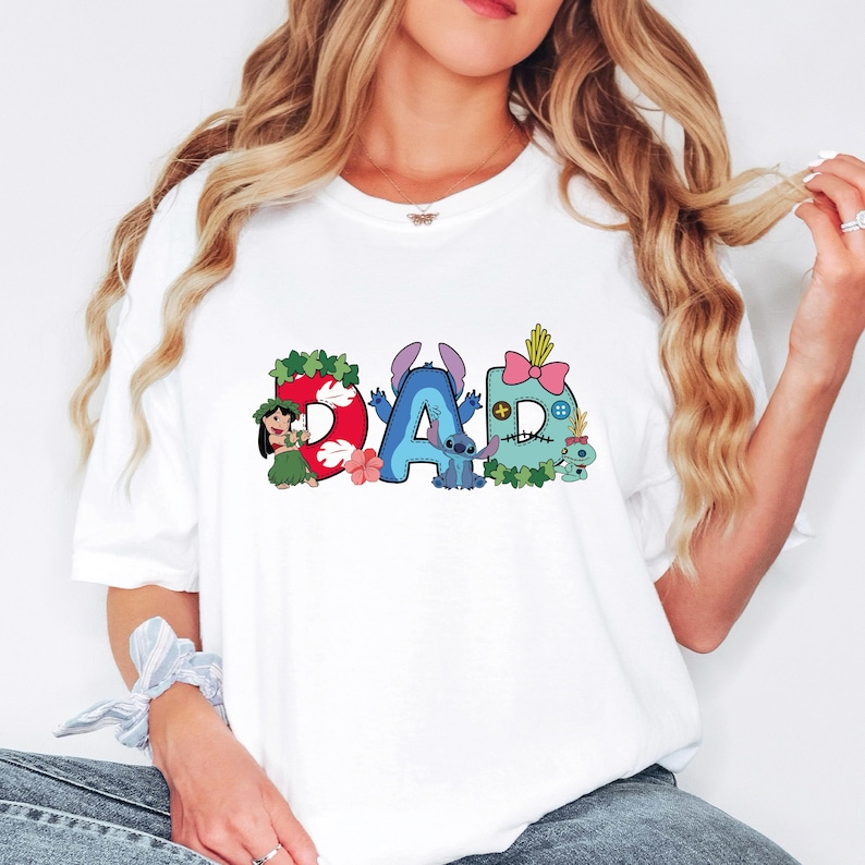 May include: A white t-shirt featuring a graphic print of the word DAD. The letter D is red with Lilo, the letter A is blue with Stitch, and the letter D is light blue with a pink bow and Scrump, all decorated with tropical floral accents.