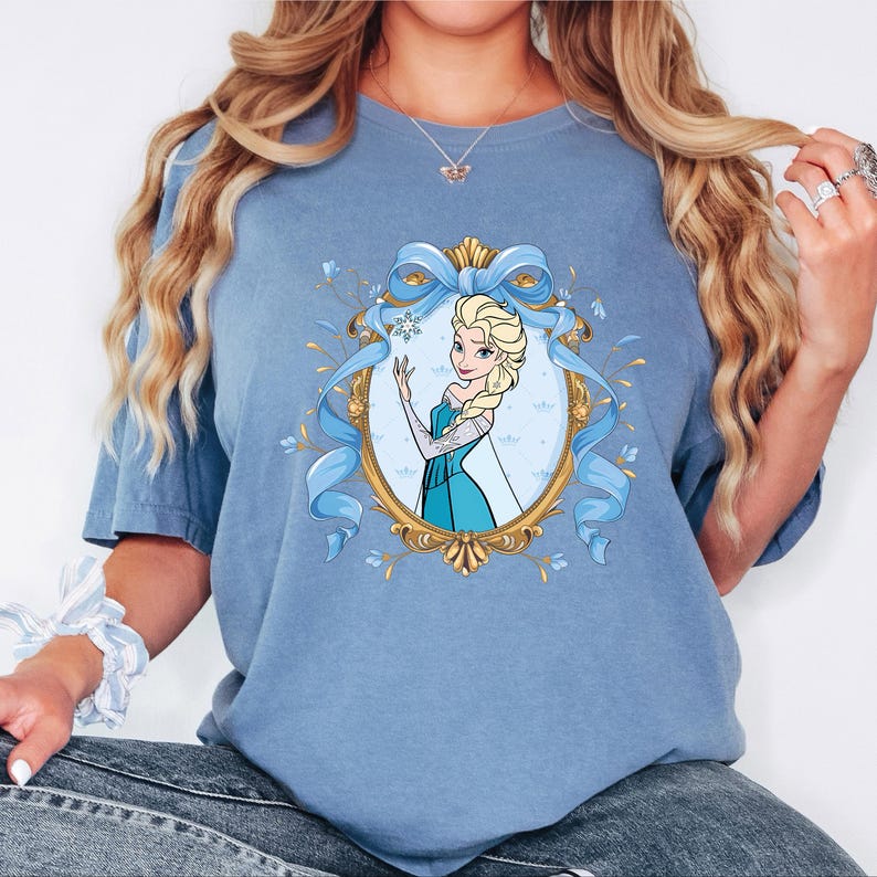 May include: A blue short-sleeve t-shirt featuring a graphic print of a blonde character in a teal dress inside an ornate gold oval frame. The frame is decorated with a large light blue ribbon bow at the top and small blue floral accents.