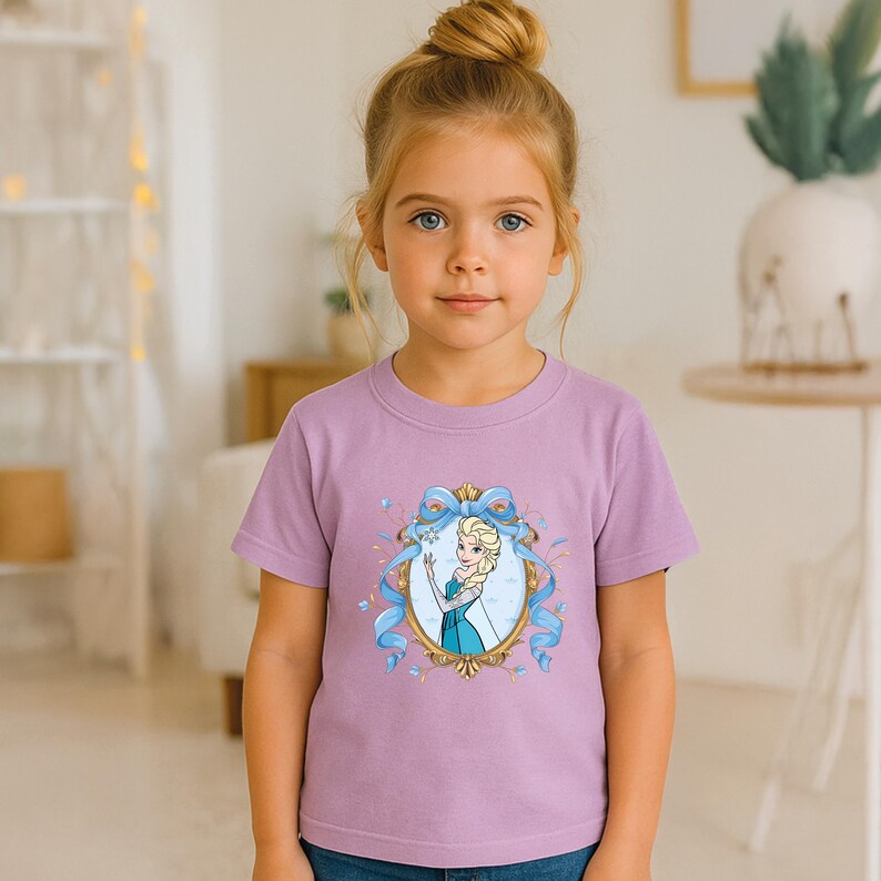 May include: A light purple short-sleeved t-shirt featuring a central graphic print of a blonde character in a blue dress inside an ornate gold oval frame, decorated with a flowing light blue ribbon and small blue snowflake accents.