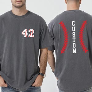 May include: Charcoal gray t-shirt with the number "42" in white and baseball stitching on the front. The back of the shirt features the word "CUSTOM" in white, with red baseball stitching accents.