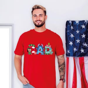 May include: A bright red short-sleeved t-shirt featuring a graphic print on the chest that spells out the word DAD. The letters are decorated with colorful cartoon characters, floral patterns, and buttons, incorporating blue, green, and pink accents.