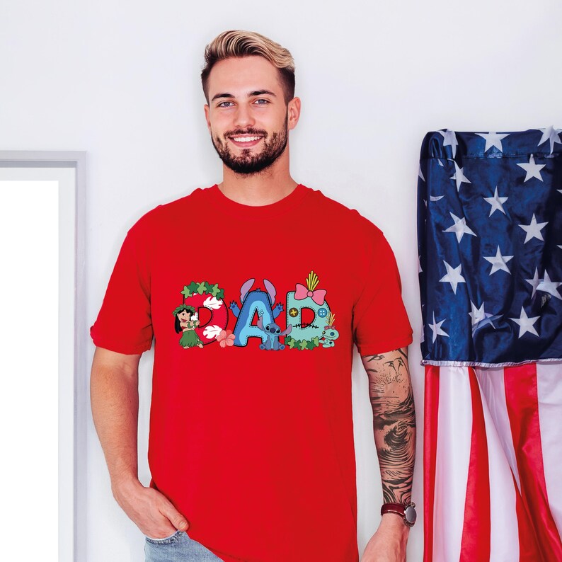 May include: A bright red short-sleeved t-shirt featuring a graphic print on the chest that spells out the word DAD. The letters are decorated with colorful cartoon characters, floral patterns, and buttons, incorporating blue, green, and pink accents.
