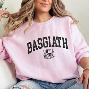 以下が含まれることがあります： 胸に太字の黒い文字で「BASGIATH」と書かれた、明るいピンク色のスウェットシャツ。その下に、盾と「SWAN COLLEGE」という言葉が入った紋章があります。スウェットシャツはカジュアルなスタイルです。