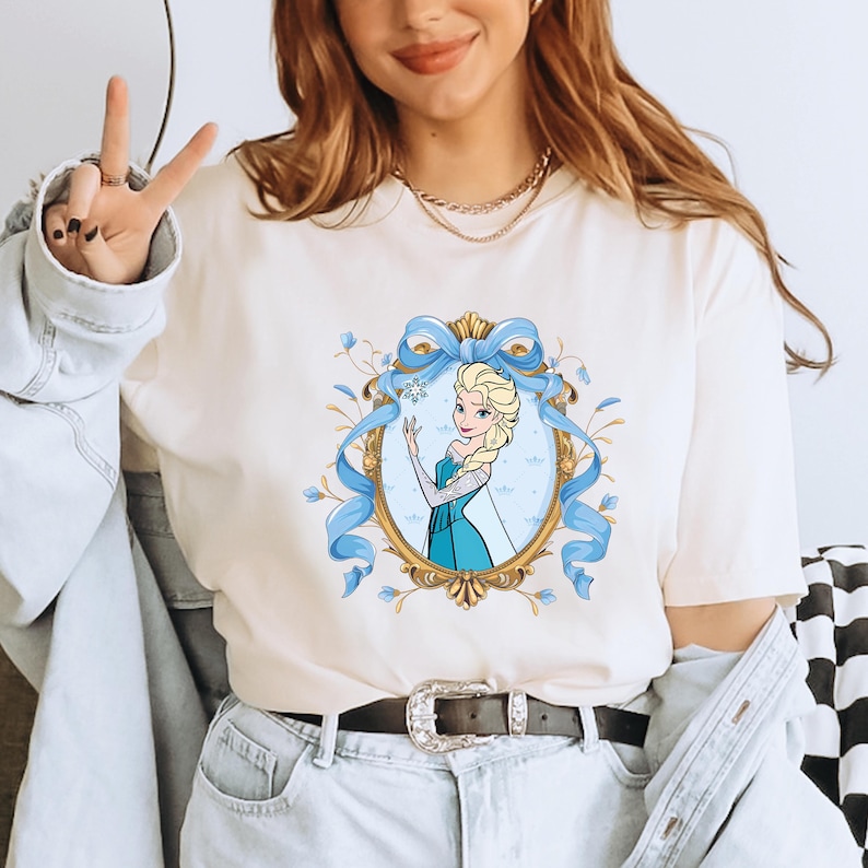 May include: A cream-colored short-sleeve t-shirt featuring a graphic print of a blonde character in a blue dress inside an ornate gold oval frame. The frame is decorated with a large light blue ribbon bow at the top and delicate floral accents.