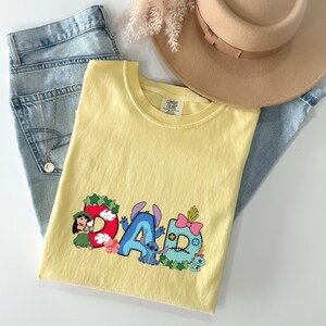 May include: A pale yellow short-sleeved t-shirt featuring a graphic print of the word DAD, where each letter is decorated with Lilo and Stitch characters, tropical flowers, and leaves. The shirt is paired with light blue denim jeans and a tan hat.