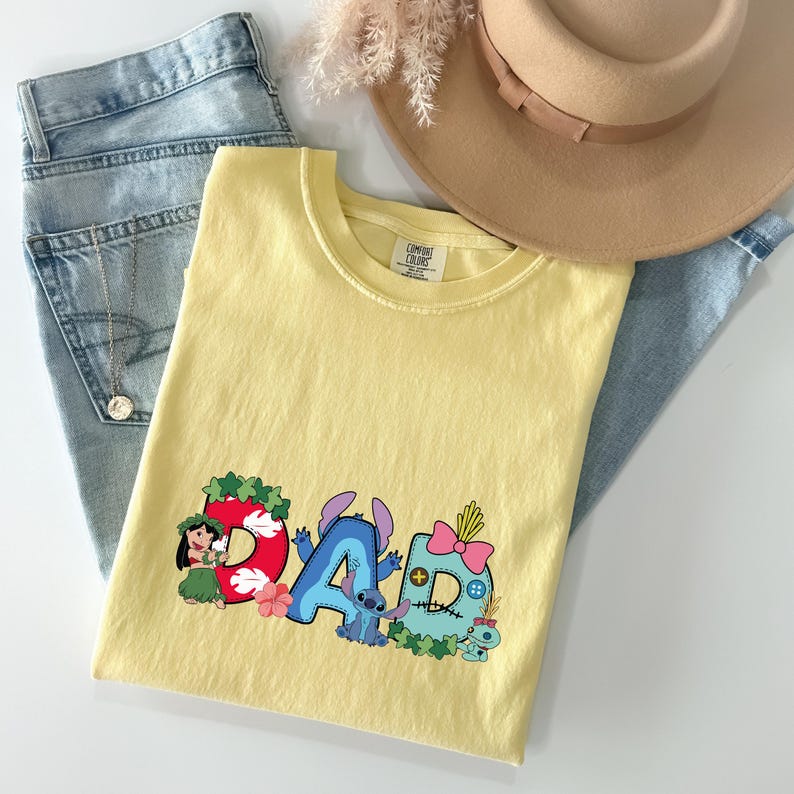 May include: A pale yellow short-sleeved t-shirt featuring a graphic print of the word DAD, where each letter is decorated with Lilo and Stitch characters, tropical flowers, and leaves. The shirt is paired with light blue denim jeans and a tan hat.