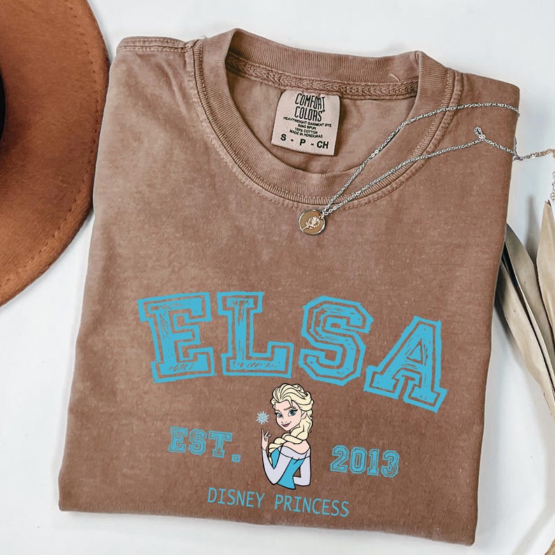 May include: A brown Comfort Colors t-shirt featuring the name "ELSA" in turquoise block letters, with an image of Elsa and the text "EST. 2013 DISNEY PRINCESS". A silver necklace with a pendant is on the shirt.