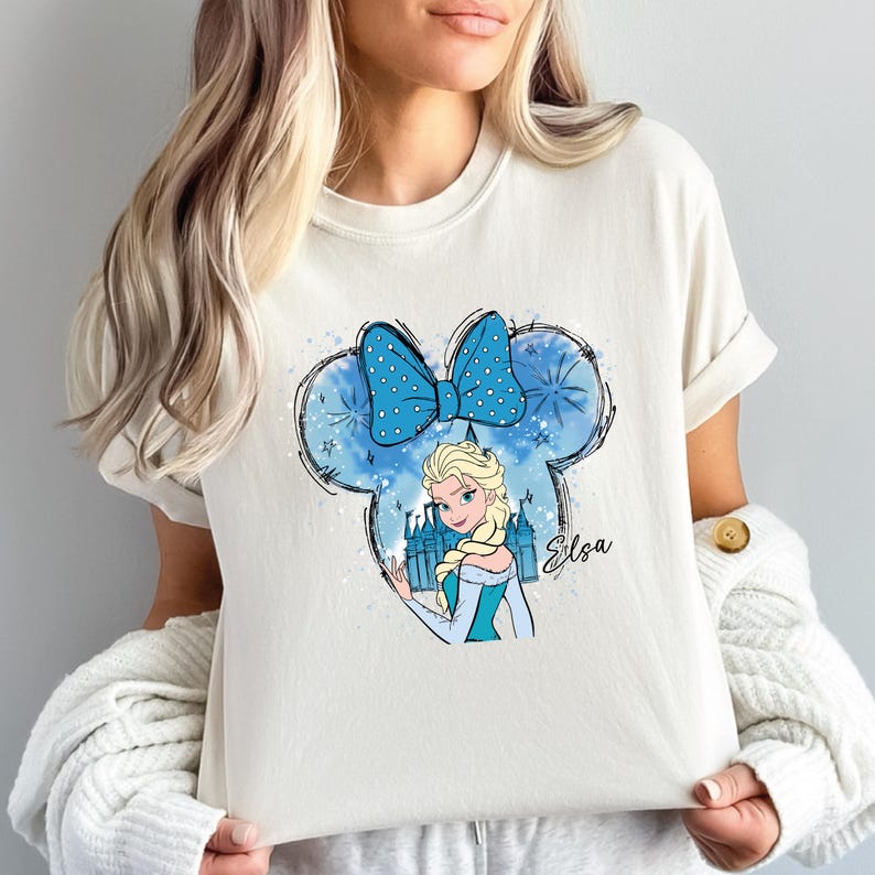 May include: Cream-colored t-shirt featuring a cartoon graphic of Elsa from Frozen. The design includes a blue polka-dot bow, a castle, and the name "Elsa" in script. The graphic is set within a Mickey Mouse ear outline.