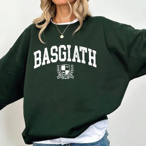 以下が含まれることがあります： 胸に「BASGIATH」の文字が大きく白くかすれた文字でプリントされた、濃い緑色のスウェットシャツ。テキストの下には紋章のデザインがあります。スウェットシャツはクルーネックで長袖です。