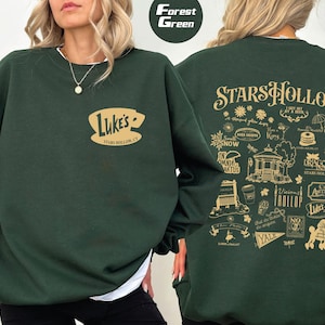 May include: Forest green sweatshirt featuring the text "Luke's STARS HOLLOW, CT" on the front and a detailed graphic on the back. The back graphic includes the text "STARS HOLLOW" and various illustrations.