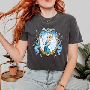 May include: A charcoal gray short-sleeve t-shirt featuring a graphic print of a blonde character in a blue dress inside an ornate gold oval frame, accented with a large light blue ribbon bow and small snowflake details centered on the chest.