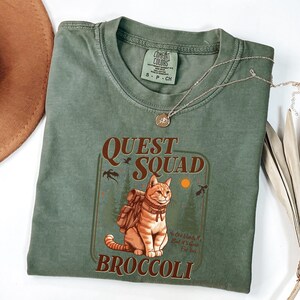 K&ouml;nnte beinhalten: Olivgr&uuml;nes T-Shirt mit einer Grafik einer Katze mit Rucksack, den Worten "Quest Squad" und "Broccoli". Das Shirt hat auch den Text "No One Hands It, But It's Good For You". Eine goldene Halskette liegt &uuml;ber dem Shirt.