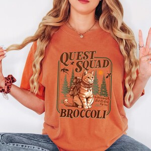 K&ouml;nnte beinhalten: Orangefarbenes T-Shirt mit einer Grafik, die eine Katze mit Rucksack, die Worte "Quest Squad" und "Broccoli" zeigt. Das Design umfasst B&auml;ume, V&ouml;gel und eine Sonne. Der Text "No One Wants It, But It's Good For You" ist ebenfalls enthalten.