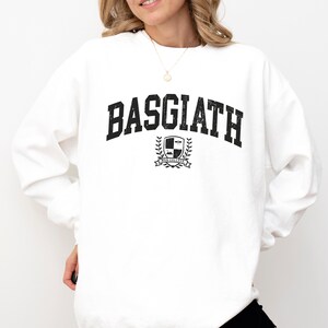 以下が含まれることがあります： 胸に「BASGIATH」の文字が大きく黒色でプリントされた白いスウェットシャツ。その下に、盾、月桂樹の葉、そして「WAR COLLEGE」の文字が入った紋章があります。長袖のスウェットシャツです。