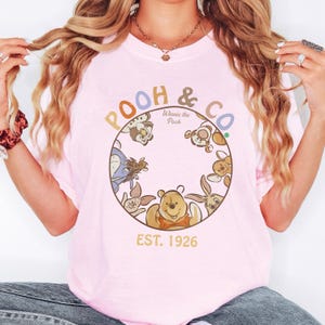 May include: A light pink t-shirt featuring a circular design with Winnie the Pooh characters. The design includes the text "POOH & CO." and "EST. 1926". The shirt has short sleeves and a crew neck.