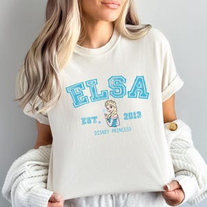 May include: Cream-colored t-shirt featuring the name "ELSA" in light blue block letters. Below the name is "EST. 2013" and a graphic of Elsa. The words "DISNEY PRINCESS" are printed below the graphic.