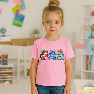 May include: A light pink short-sleeved t-shirt featuring a graphic print of the word DAD. The letters are decorated with cartoon characters including Lilo and Stitch, with floral accents, a pink bow, and various colorful patterns on each letter.