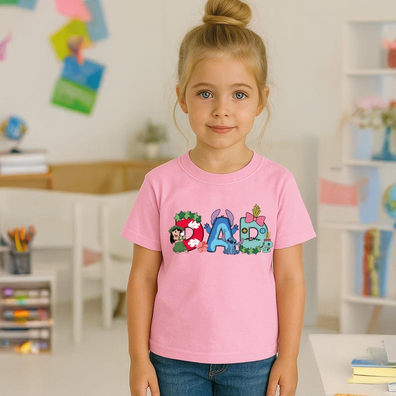 May include: A light pink short-sleeved t-shirt featuring a graphic print of the word DAD. The letters are decorated with cartoon characters including Lilo and Stitch, with floral accents, a pink bow, and various colorful patterns on each letter.