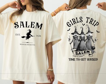 May include: Cream-colored t-shirt with black graphics. The front features "SALEM" above a witch on a broom, with "EST. 1626" and "MASSACHUSETTS" below. The back displays "GIRLS TRIP" and three witches with the text "TIME TO GET WICKED".
