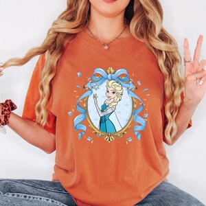 May include: A burnt orange t-shirt featuring a central graphic of a blonde character in a blue dress, framed by an ornate gold oval border and a light blue ribbon. The character has long blonde hair and is wearing a blue gown with sheer sleeves.