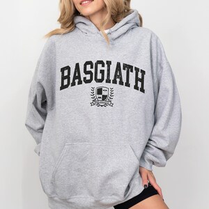以下が含まれることがあります： 胸に「BASGIATH」の文字が大きく黒色でプリントされたライトグレーのパーカー。その下に、盾と「WAR COLLEGE」の文字が入った紋章があります。パーカーにはフロントポケットとフードが付いています。