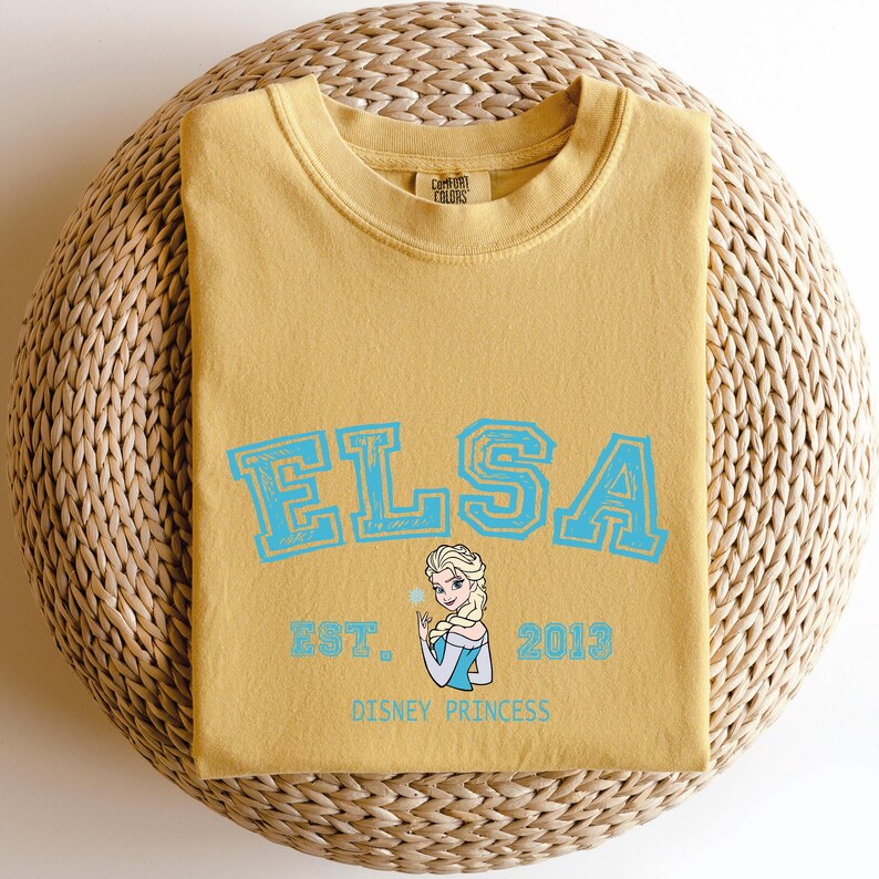 May include: Mustard yellow t-shirt with the name "ELSA" in light blue block letters. Below the name is a graphic of Elsa, the Disney princess, with the text "EST. 2013 DISNEY PRINCESS". The shirt is folded on a woven, circular surface.
