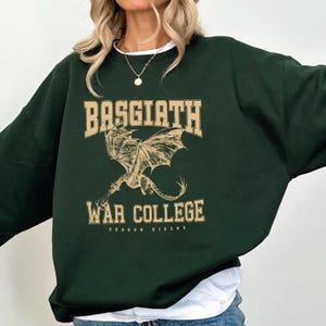 May include: Dark green sweatshirt with the text "BASGIATH WAR COLLEGE" in a tan, collegiate-style font. A tan dragon graphic is centered below the text. The words "DRAGON RIDERS" are below the graphic.