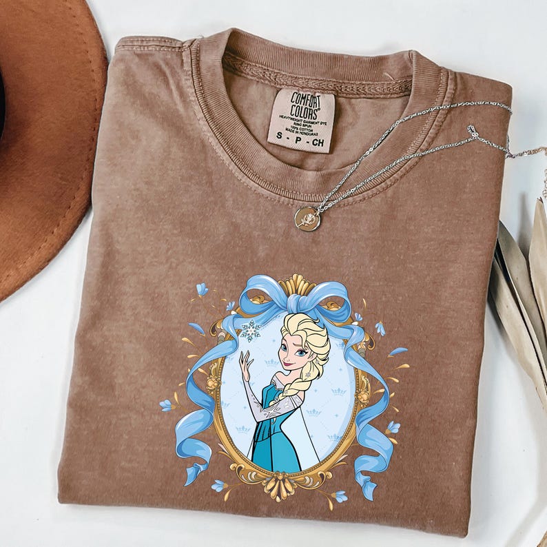 May include: A brown short-sleeve t-shirt featuring a graphic of a blonde character in a blue dress inside an ornate gold oval frame with a blue ribbon. The shirt has a Comfort Colors label at the neck and a silver-toned necklace resting on top.
