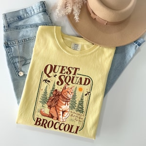 K&ouml;nnte beinhalten: Hellgelbes T-Shirt mit einer Grafik einer Katze mit Rucksack, den Worten "QUEST SQUAD" und "BROCCOLI". Das Shirt wird mit hellblauen Jeans und einem beigen Hut kombiniert.