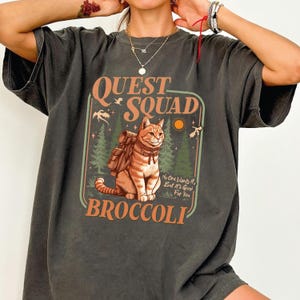 K&ouml;nnte beinhalten: Dunkelgraues T-Shirt mit einer Grafik, die eine Katze mit Rucksack, die Worte "QUEST SQUAD" und "BROCCOLI" zeigt. Das Design beinhaltet eine Waldszene und den Text "No One Wants It, But It's Good For You."