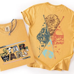 K&ouml;nnte beinhalten: Senfgelbe T-Shirts mit Star Wars-Grafiken. Ein T-Shirt zeigt den Text "STAR WARS" mit Charakterillustrationen und Lichtschwertern. Das andere Shirt zeigt Darth Vader, einen Sturmtruppler und den Todesstern.