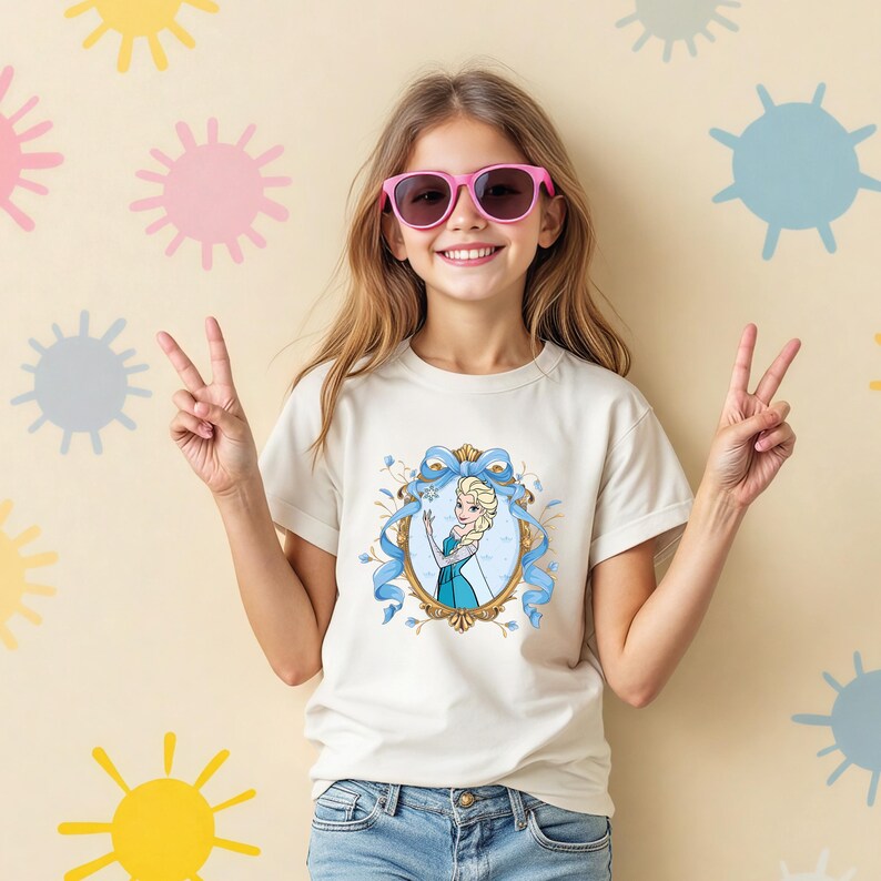 May include: A short-sleeved cream-colored t-shirt featuring a graphic print of a blonde character in a blue dress inside an ornate gold oval frame with a blue ribbon bow. The shirt is paired with blue denim jeans and pink-framed sunglasses.