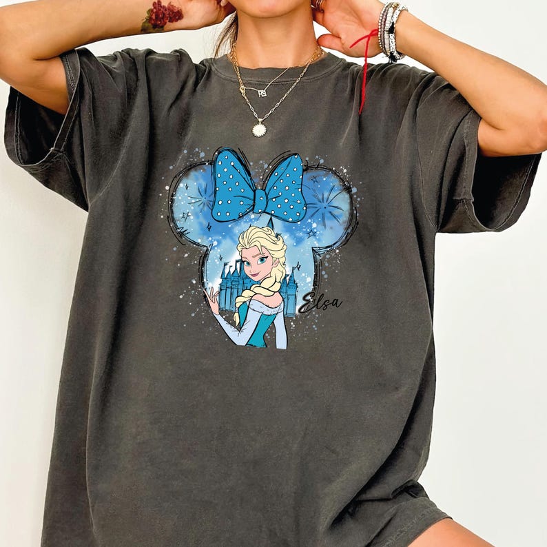 May include: Dark gray t-shirt featuring a graphic of Elsa from Frozen inside a Mickey Mouse silhouette. The design includes a blue bow with white polka dots, a castle, and the name "Elsa" in cursive.