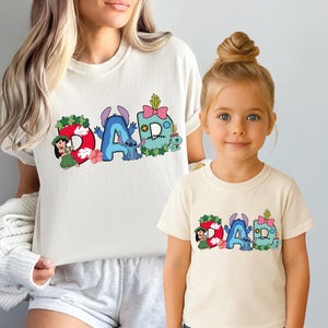May include: Two cream-colored short-sleeve t-shirts featuring a graphic print of the word DAD. The letters are decorated with Lilo and Stitch characters, tropical leaves, flowers, and a pink bow. The design includes blue, red, green, and pink colors.