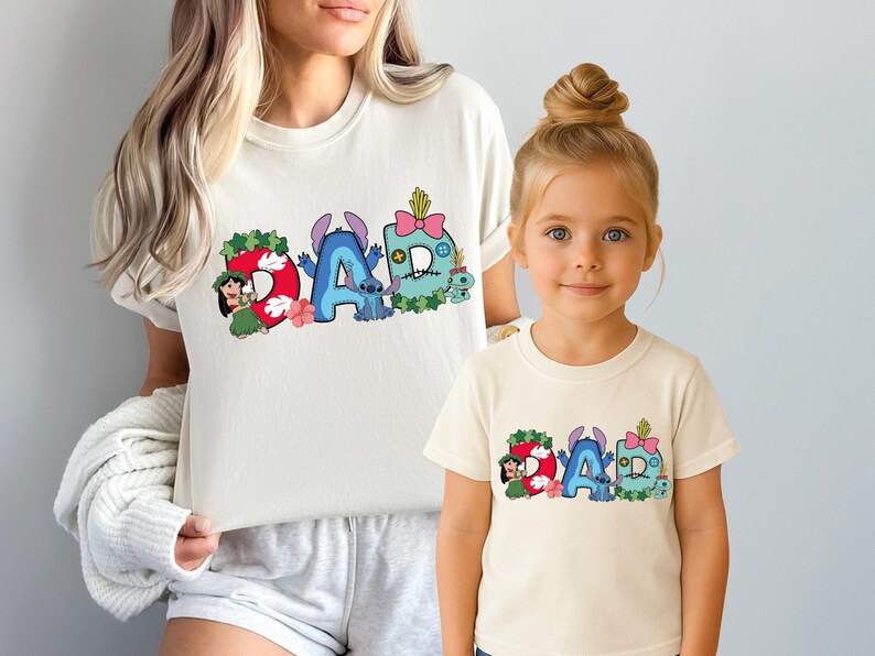 May include: Two cream-colored short-sleeve t-shirts featuring a graphic print of the word DAD. The letters are decorated with Lilo and Stitch characters, tropical leaves, flowers, and a pink bow. The design includes blue, red, green, and pink colors.