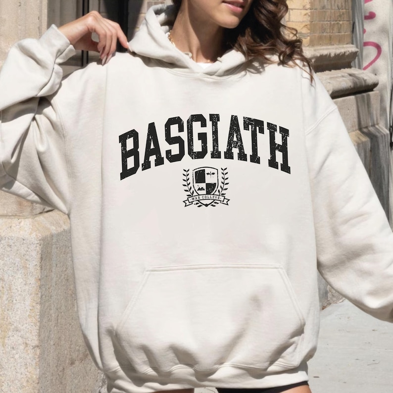 以下が含まれることがあります： 胸に「BASGIATH」の文字が太字の黒色でかすれたようにプリントされたクリーム色のパーカー。テキストの下には、盾、月桂樹の葉、そして「WAR COLLEGE」の文字が入った紋章があります。パーカーには、ドローストリングフードとフロントポケットが付いています。