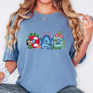 May include: A blue short-sleeve t-shirt featuring a graphic print of the word DAD. The letter D is red with Lilo, the letter A is blue with Stitch, and the letter D is light blue with a pink bow, buttons, and a small green creature.