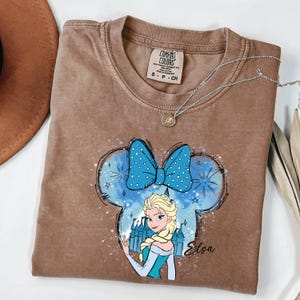 May include: Brown Comfort Colors t-shirt featuring a graphic of Elsa from Frozen inside a Mickey Mouse-shaped design with a blue bow. The design includes a castle and the name "Elsa". A silver necklace with a pendant is on the shirt.