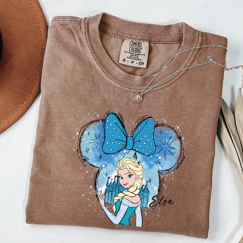 May include: Brown Comfort Colors t-shirt featuring a graphic of Elsa from Frozen inside a Mickey Mouse-shaped design with a blue bow. The design includes a castle and the name "Elsa". A silver necklace with a pendant is on the shirt.