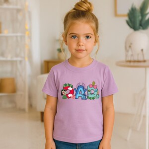 May include: A light purple short-sleeved t-shirt featuring a graphic print of the word DAD. The letters are decorated with colorful cartoon characters, including a girl in a green dress, a blue alien, and various floral and tropical motifs.