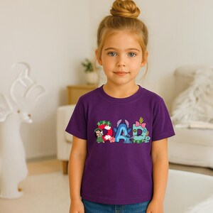 May include: A purple short-sleeved t-shirt featuring the word DAD printed in large, colorful letters. Each letter is decorated with cartoon characters and floral motifs, including a hula dancer, a blue alien, and a pink bow on a green background.