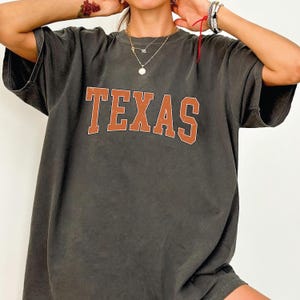 May include: Dark gray oversized t-shirt with the word "TEXAS" in large, orange, collegiate-style lettering. The shirt has a vintage, worn look.