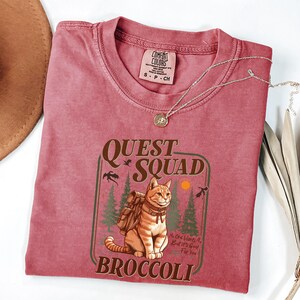 K&ouml;nnte beinhalten: Ein staubrosa Comfort Colors T-Shirt mit einer Grafik einer orangefarbenen Tabby-Katze mit Rucksack, den Worten "QUEST SQUAD" und "BROCCOLI". Eine silberne Halskette mit Anh&auml;nger liegt auf dem Shirt. Das Shirt hat die Gr&ouml;&szlig;e S-CH.