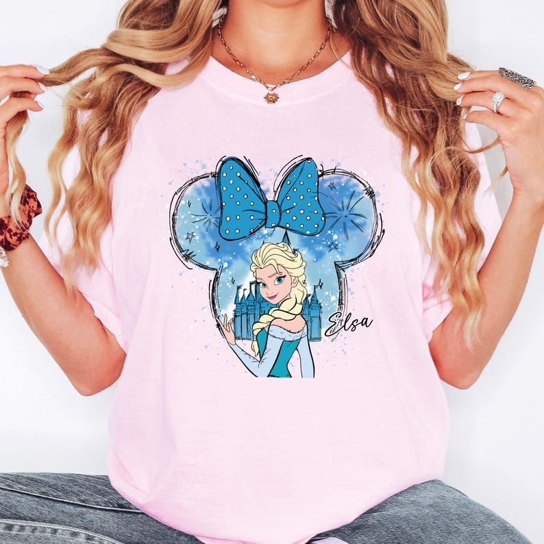 May include: A light pink t-shirt featuring a cartoon illustration of Elsa from Frozen. The design includes a blue bow, castle, and the name "Elsa". The shirt is a crew neck style.