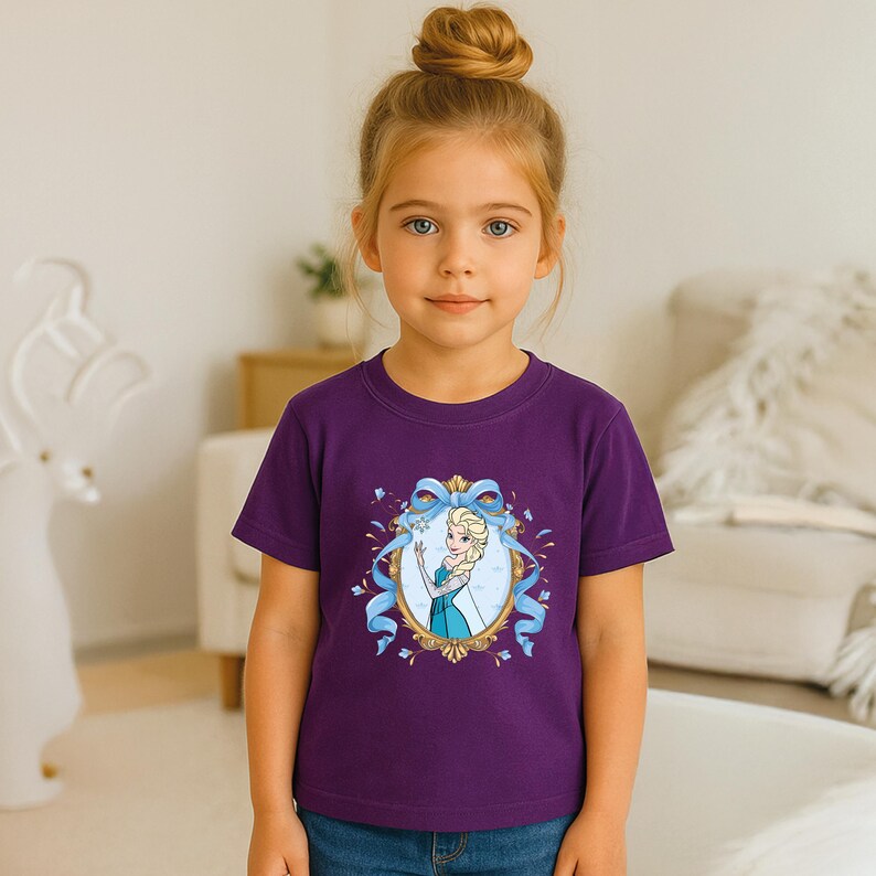 May include: A purple short-sleeved t-shirt featuring a central graphic of a blonde character in a blue dress inside an ornate gold oval frame, accented with a light blue ribbon bow at the top and small blue sparkles surrounding the frame.